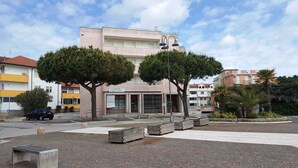 A building with a sign for 'BANCA DI PIAZZA' and a 'HOTEL' sign nearby. There are trees in front of the building, benches, and a paved area.