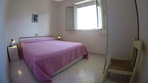 A small, neatly arranged bedroom with a single bed, a chair, a window with curtains, and a ceiling light.