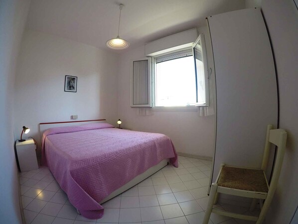 A small, neatly arranged bedroom with a single bed, a chair, a window with curtains, and a ceiling light.