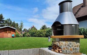 An outdoor stone and metal barbecue grill with a chimney, set in a grassy yard with a wooden cabin and trees in the background.