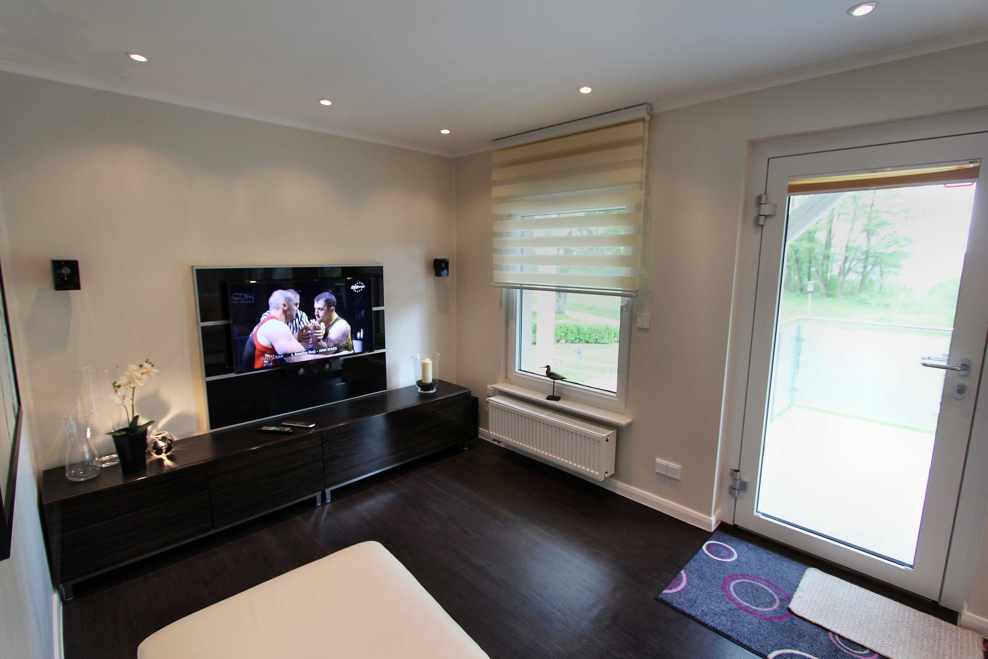 A modern living room with a flat-screen TV, dark wood furniture, a large window with blinds, and a door leading outside.