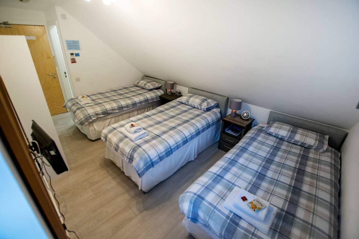 A room with three beds, each with a plaid blanket and pillows, a bedside table with a lamp, and a door with a sign on the wall.