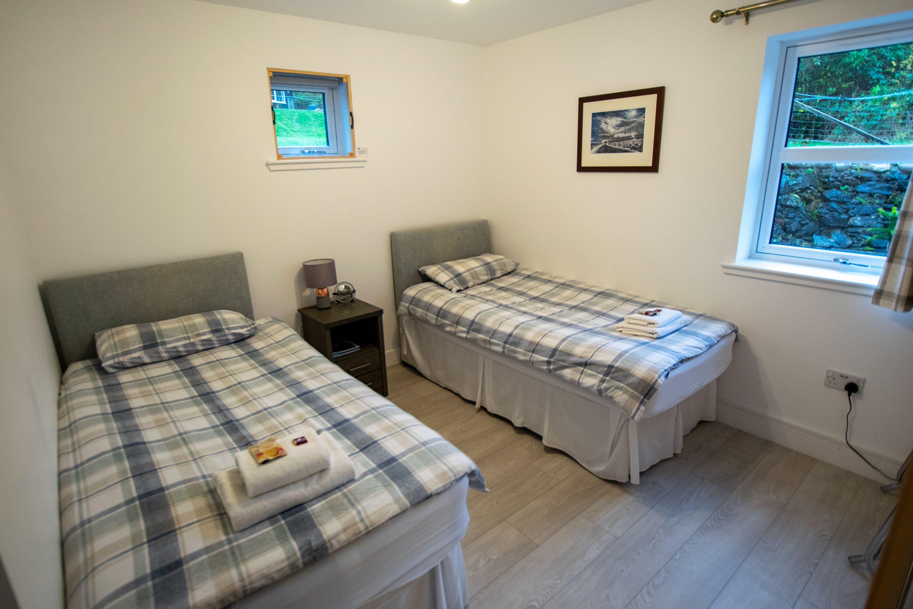 A bedroom with two beds, a small window, a nightstand, and a wall-mounted picture.