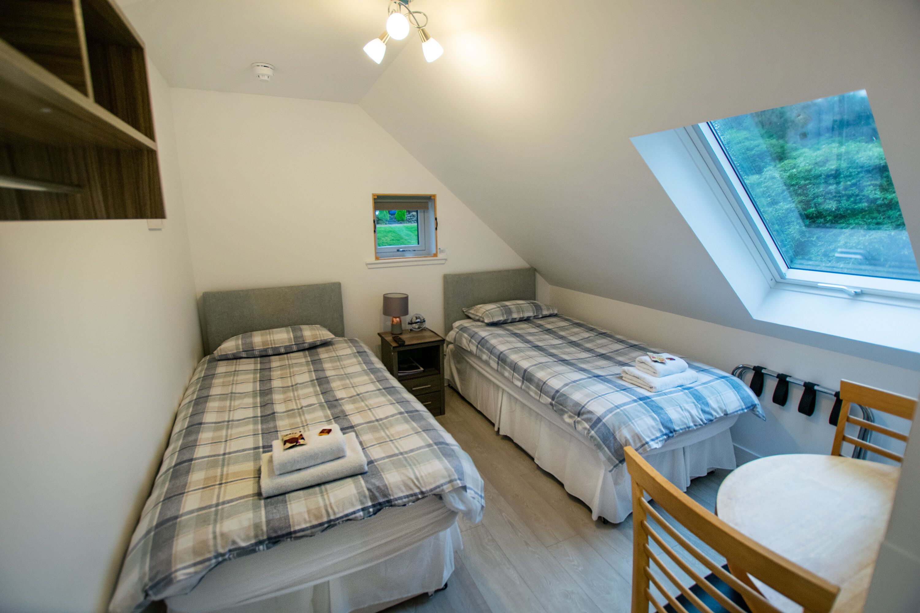 Une chambre de dortoir avec deux lits, une petite fenêtre, un luminaire au plafond et une chaise en bois.