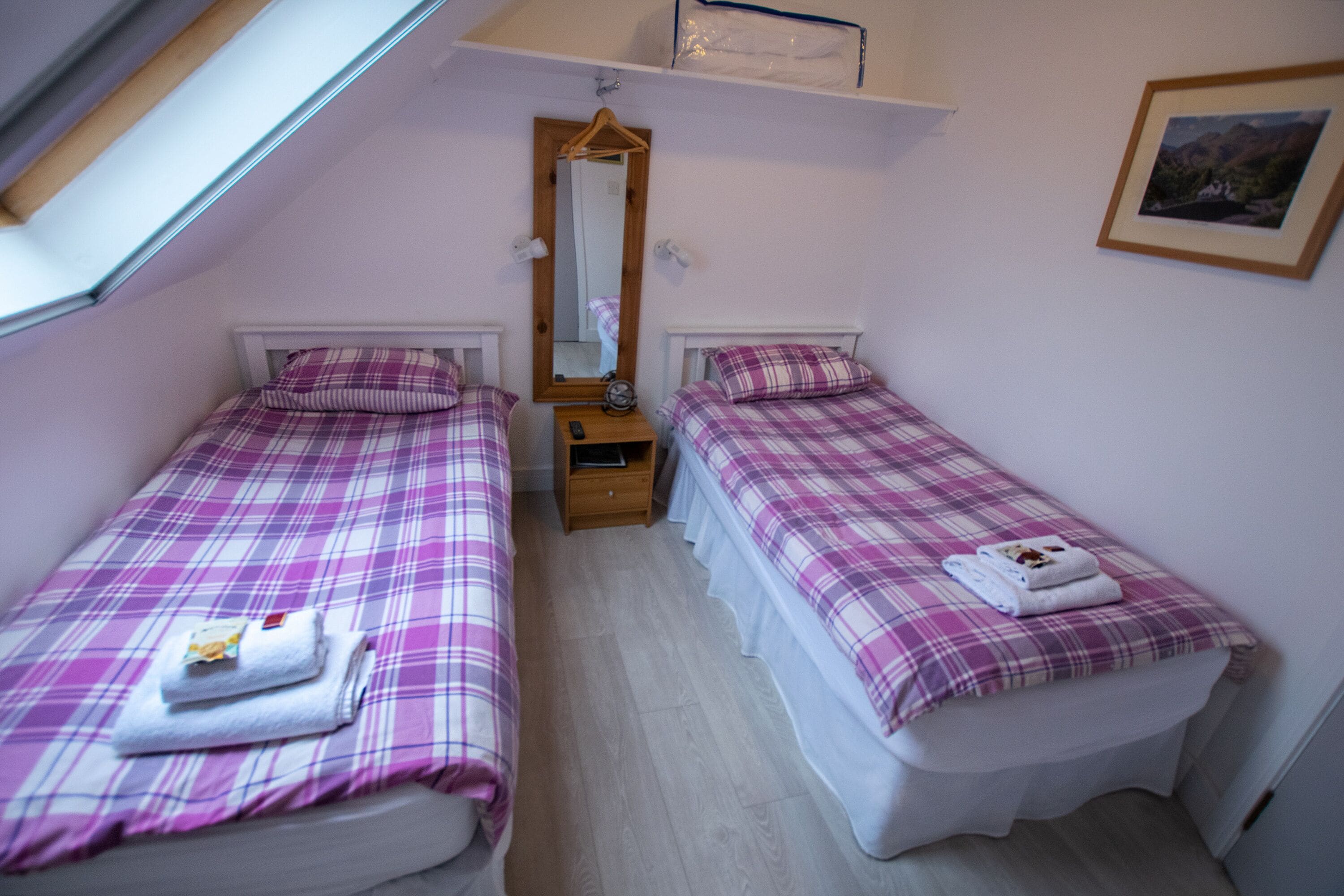 Un dormitorio con dos camas, cada una con cubrecama y almohadas a cuadros. Entre las camas hay una mesita de noche de madera, un espejo sobre la mesita y un cuadro en la pared.