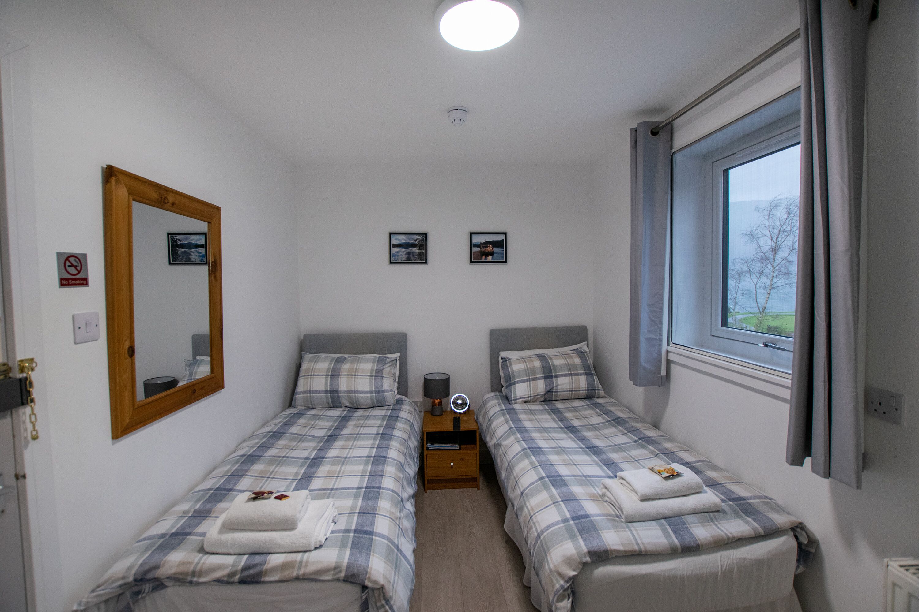 A twin room with two beds, a mirror, a bedside table with a lamp, a window with curtains, and a view of the outdoors.