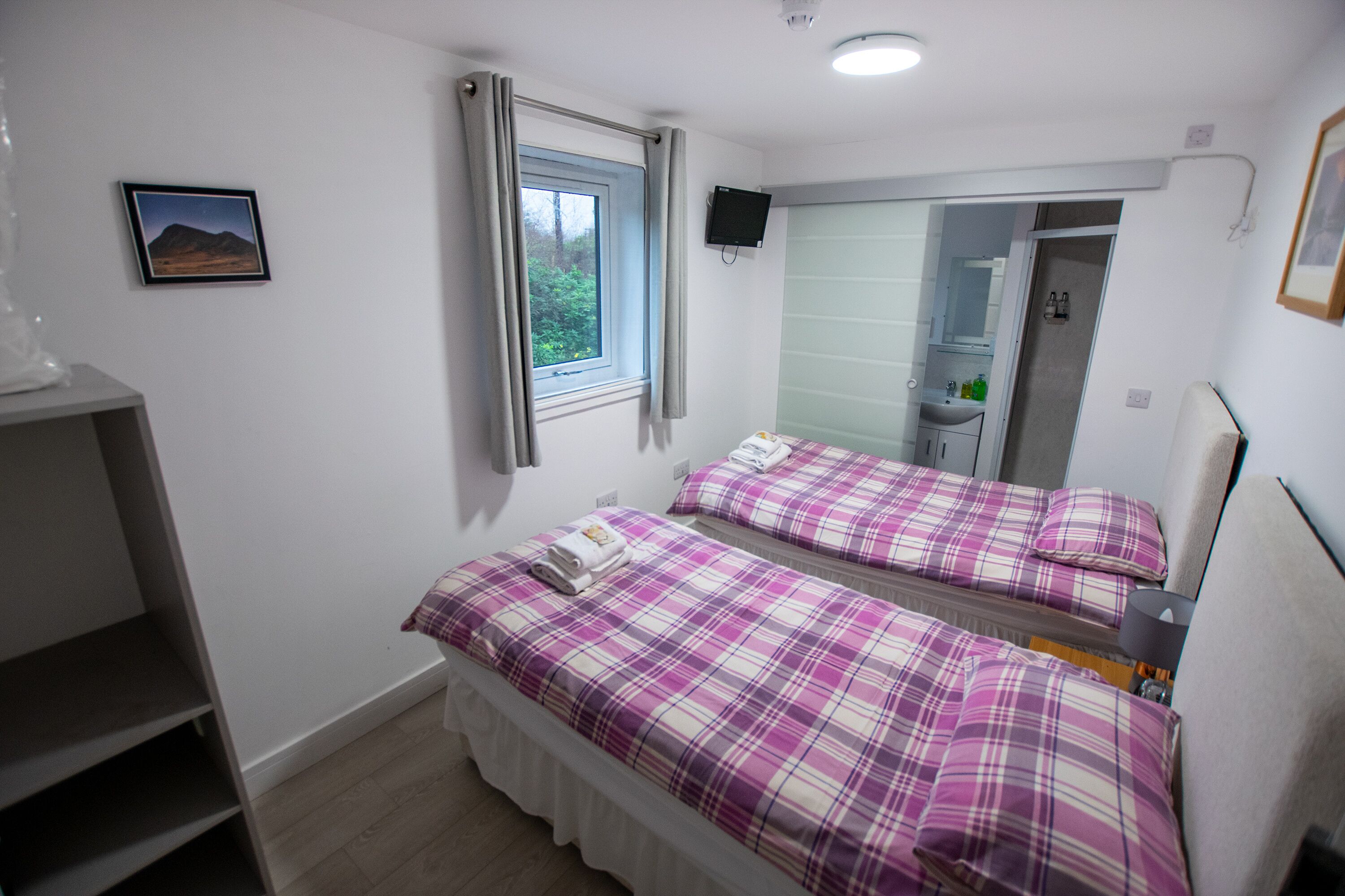 A bedroom with two beds, a window, a mirror, a TV, and a bathroom visible through the mirror.