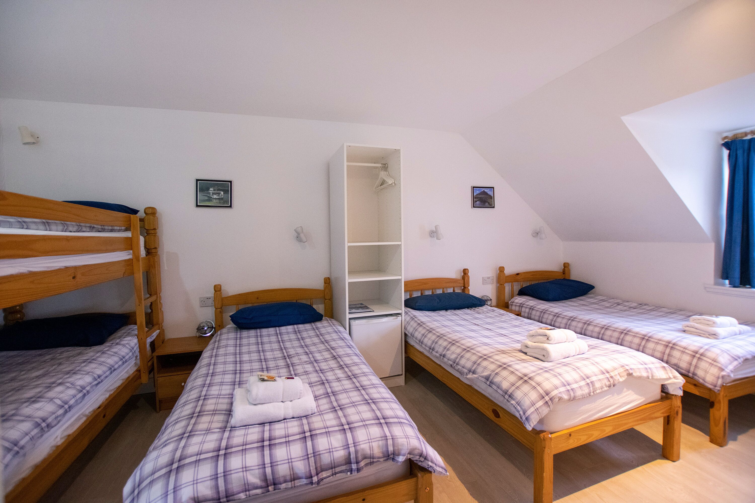 Un dormitorio con cuatro literas de madera, cada una con una manta y almohadas de cuadros. Se observa un armario blanco en el centro y un escritorio pequeño con una lámpara.