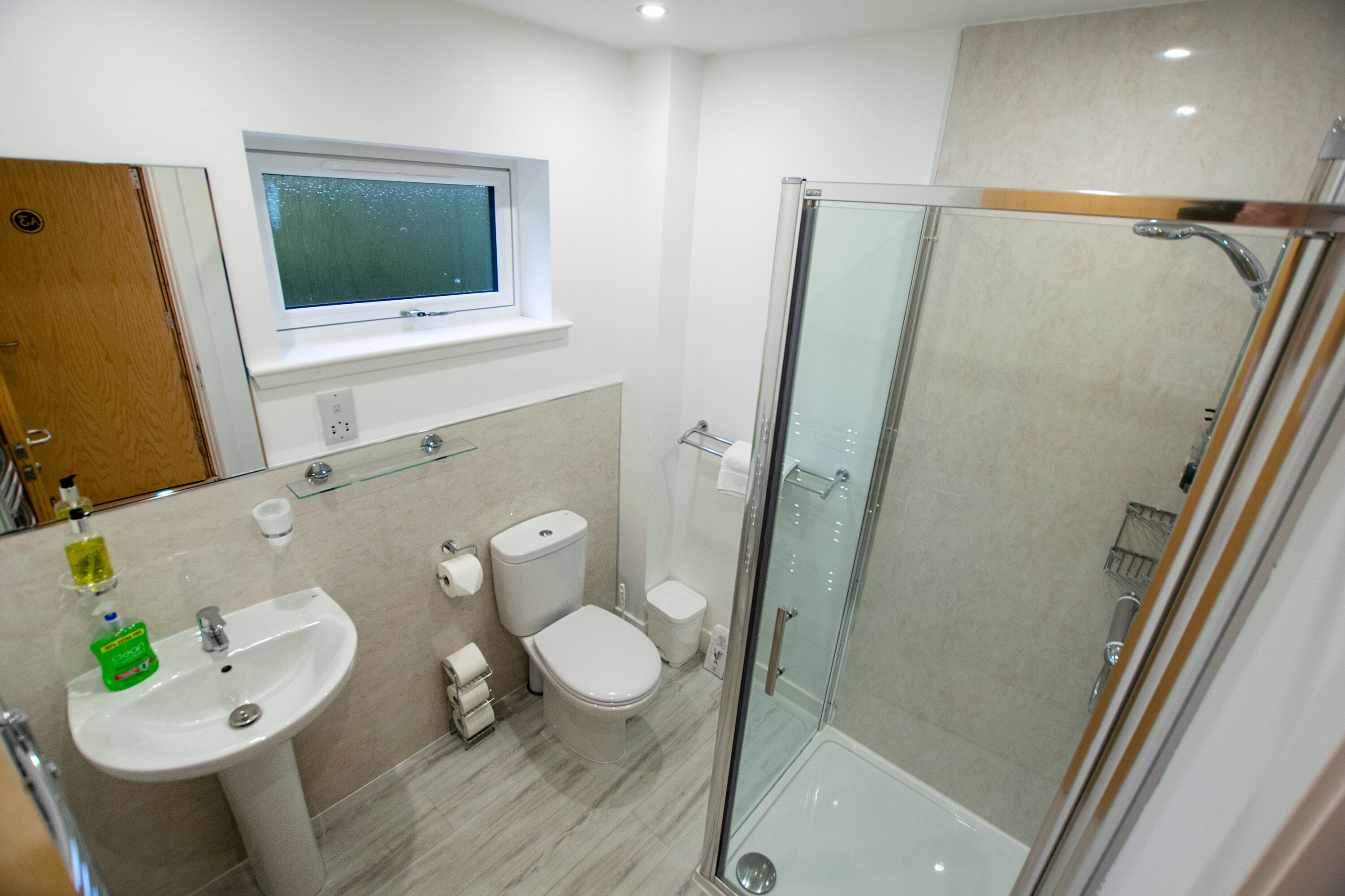 Een moderne badkamer met een douche met glazen wand, een wastafel, een toilet en een raam boven de wastafel.