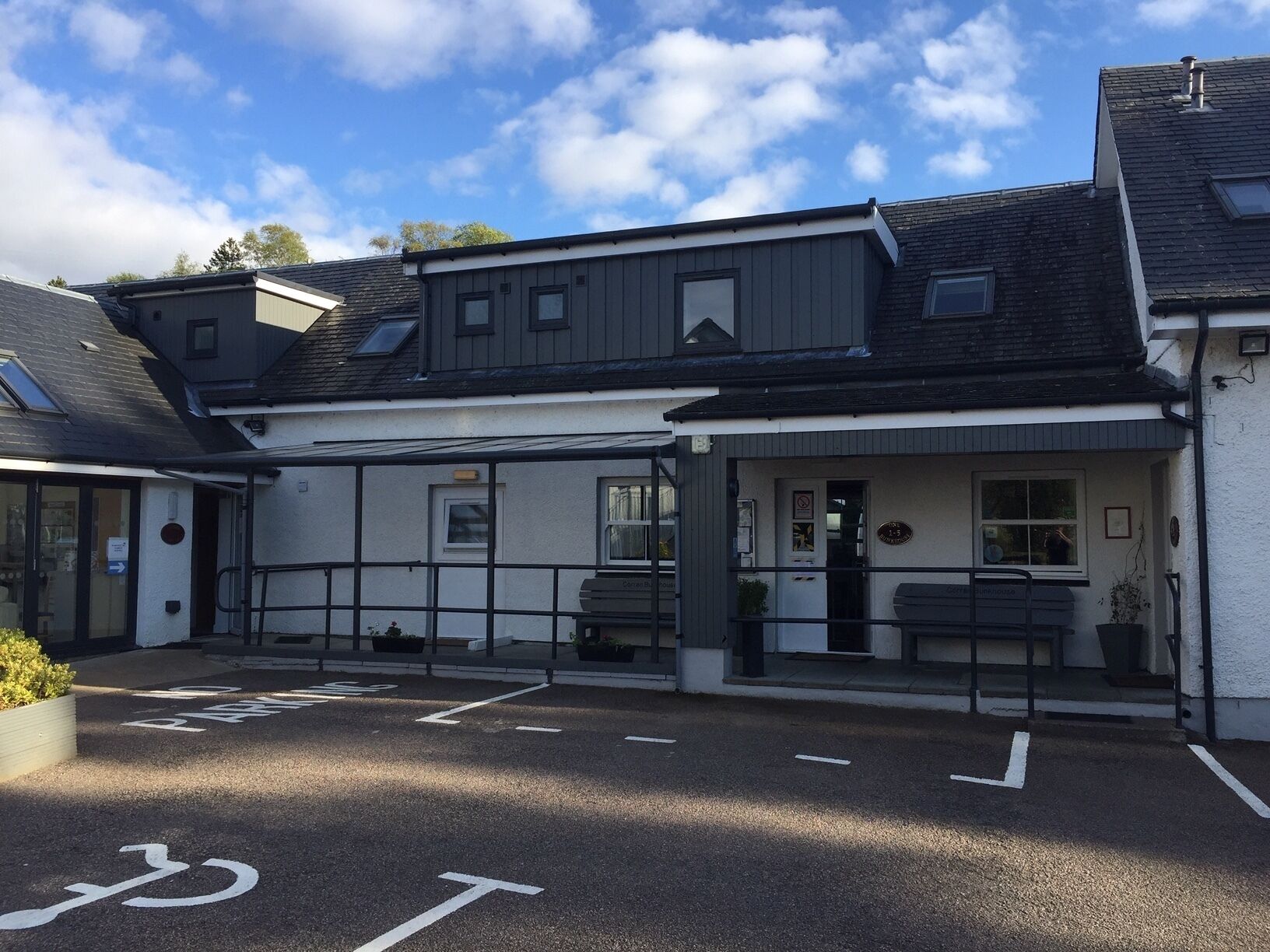 A two-story building with a flat roof, a front entrance with a door, and a small porch area with benches. There are parking spaces marked with a bicycle symbol and directional arrows.