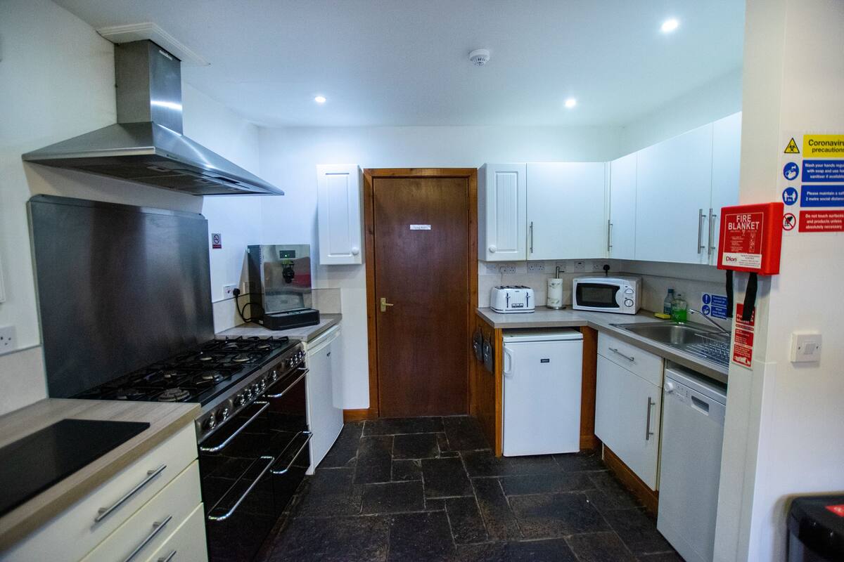 A compact kitchen with a stove, microwave, and toaster. There is a fire blanket and a sign about coronavirus precautions.