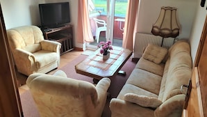 A living room with a sofa, armchairs, a TV, a coffee table with flowers, and a view of the outdoors through a sliding door.