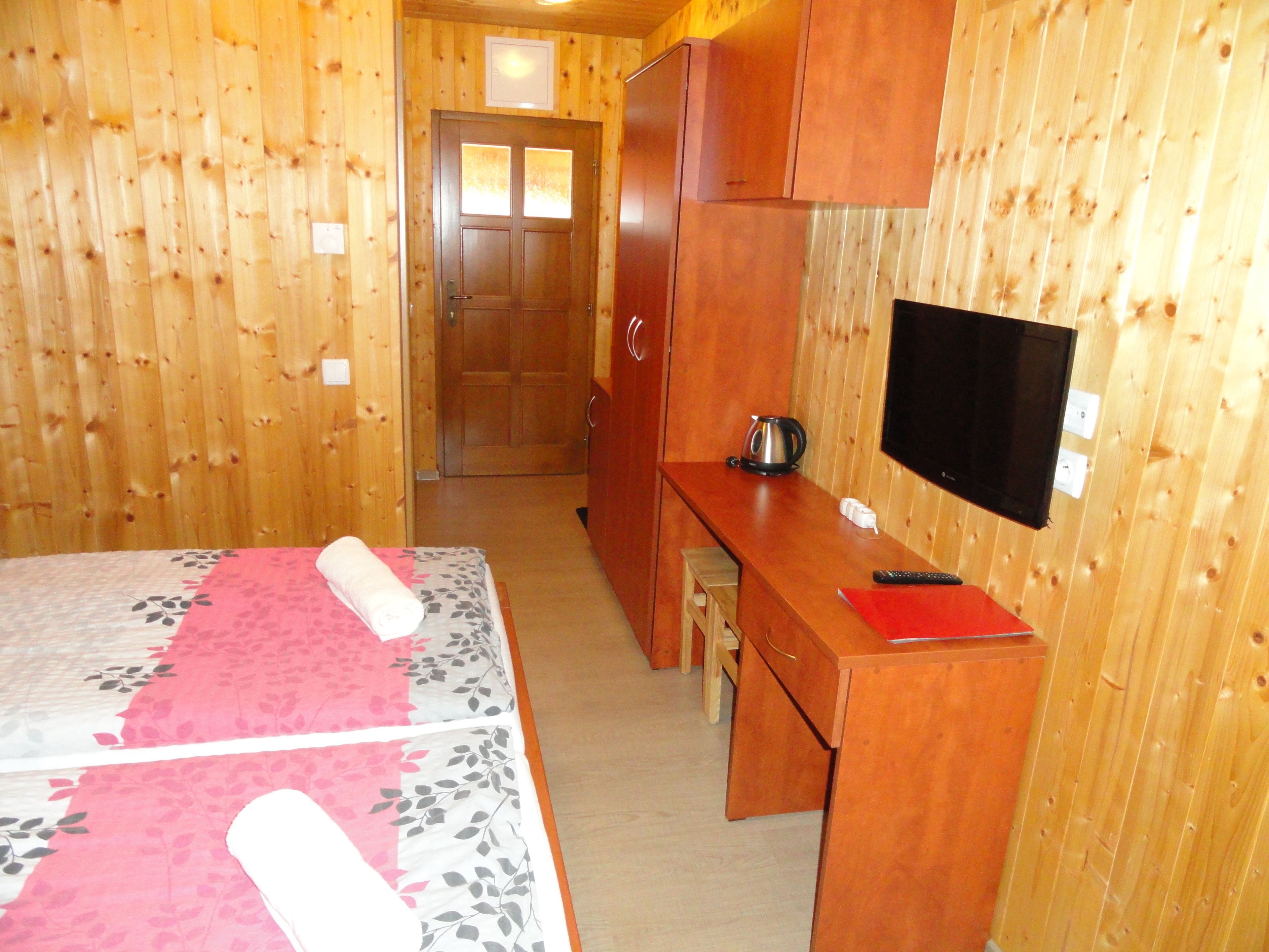 Ein Zimmer mit Holzwänden, einem Bett, einem Schreibtisch, einem Fernseher und einer Tür zu einem anderen Raum.