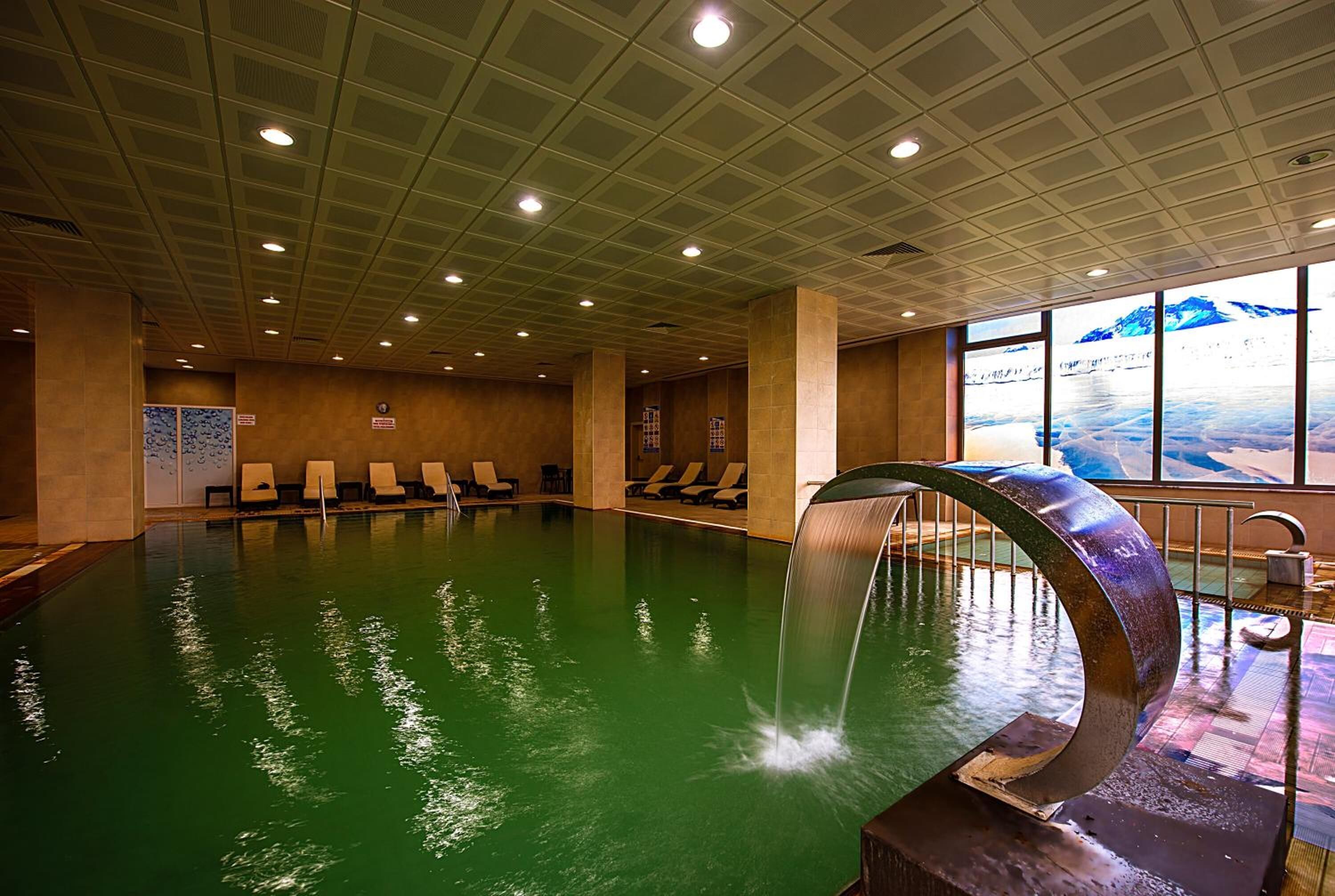 An indoor swimming pool with a waterfall feature, lounge chairs, and a view of a snowy landscape through large windows.