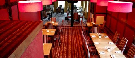A dining area with red walls, striped chairs, and a patterned carpet. Tables are set with white tablecloths and centerpieces.