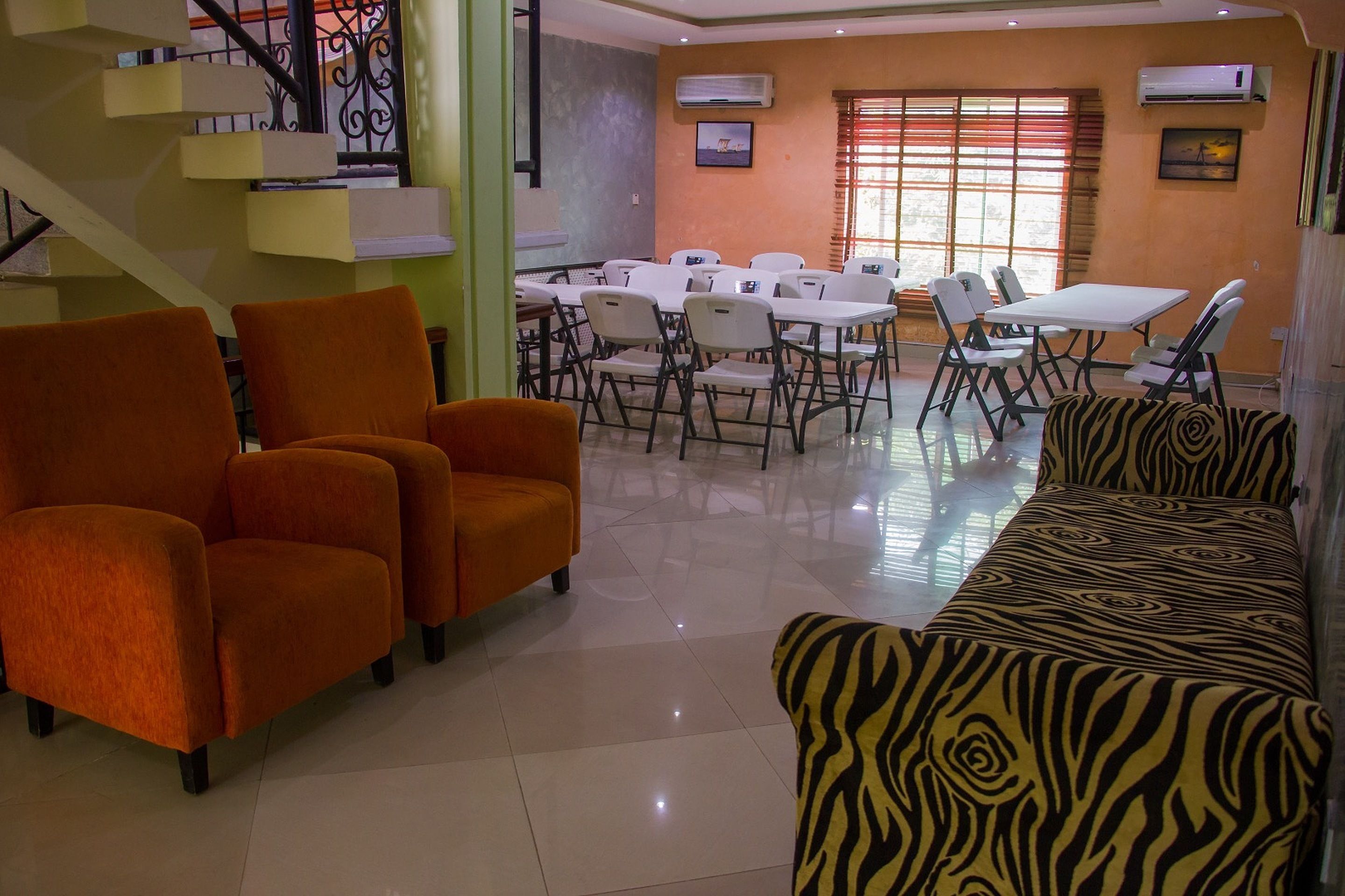 A hotel lobby with orange armchairs, a zebra-patterned sofa, a dining area with tables and chairs, and a staircase with a metal railing.