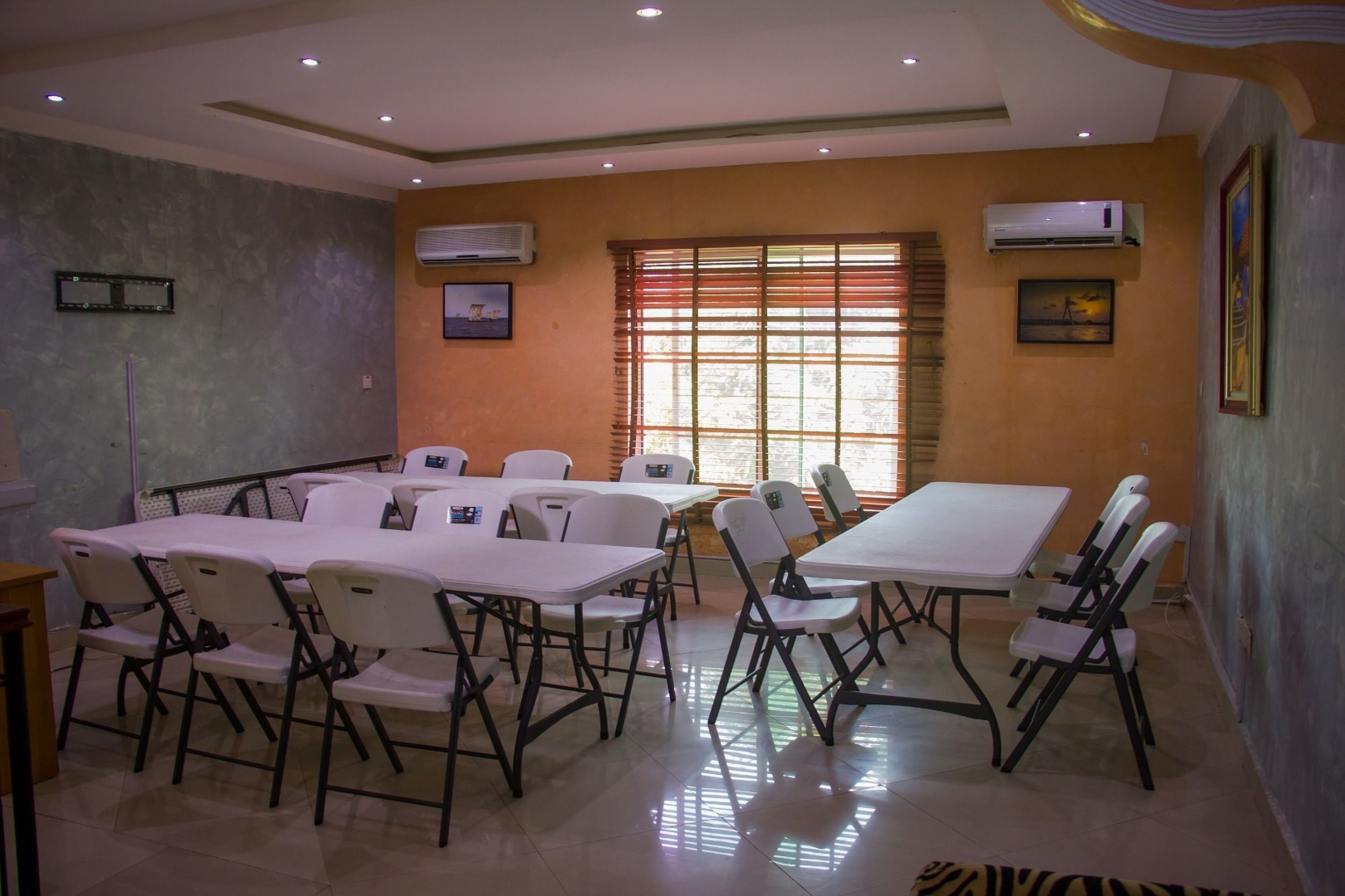 A conference room with white tables and chairs, air conditioning units, and a window with blinds.