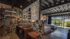 A room with a unique geometric tile pattern on the walls, a wooden bench, a bed with a view through large windows, and a small table with a bucket.