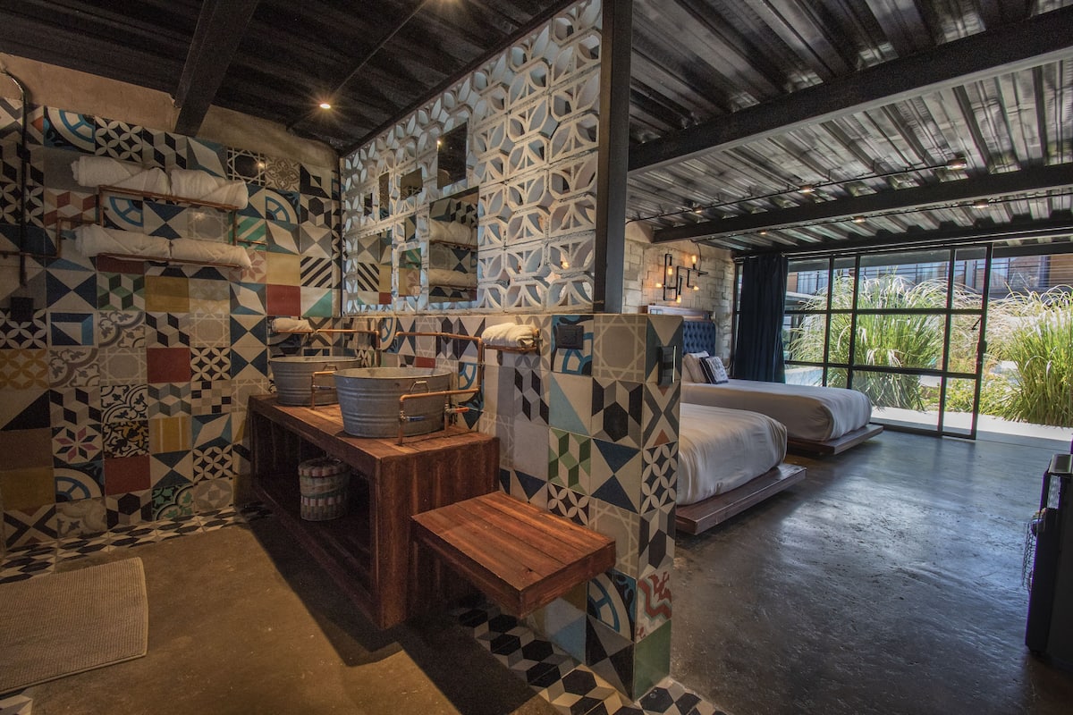 A room with a unique geometric tile pattern on the walls, a wooden bench, a bed with a view through large windows, and a small table with a bucket.