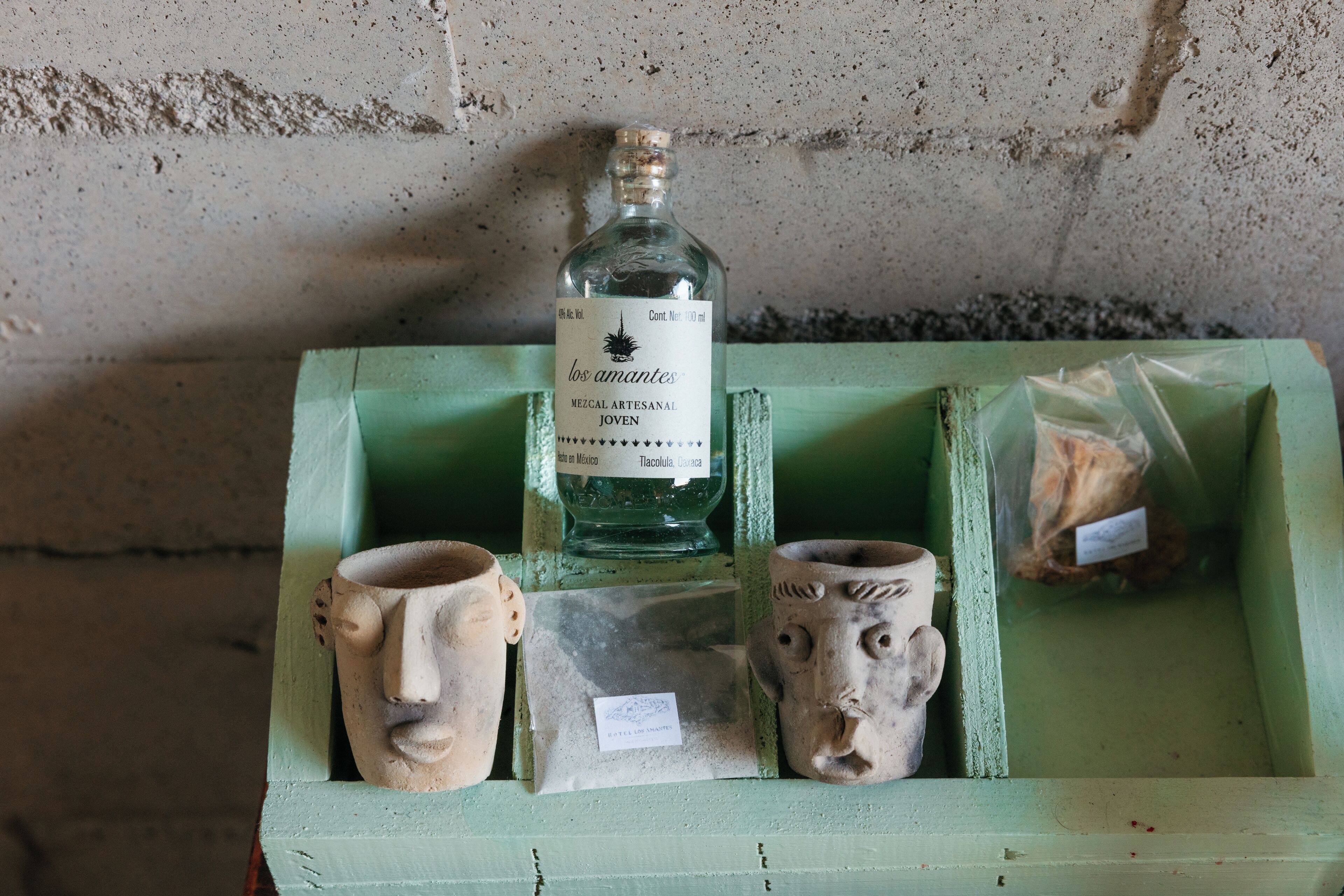 A display of ceramic items and a bottle of mezcal on a green wooden shelf against a concrete wall.