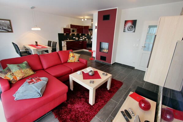 Un salon moderne avec un canapé rouge, une table basse et un tapis rouge. On aperçoit au fond une cuisine avec des meubles rouges et un coin repas.