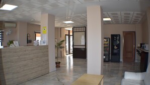 A reception area with a reception desk, a potted plant, a wooden table, a white sofa, and a refrigerator. - Hostal Alamare (Seville)
