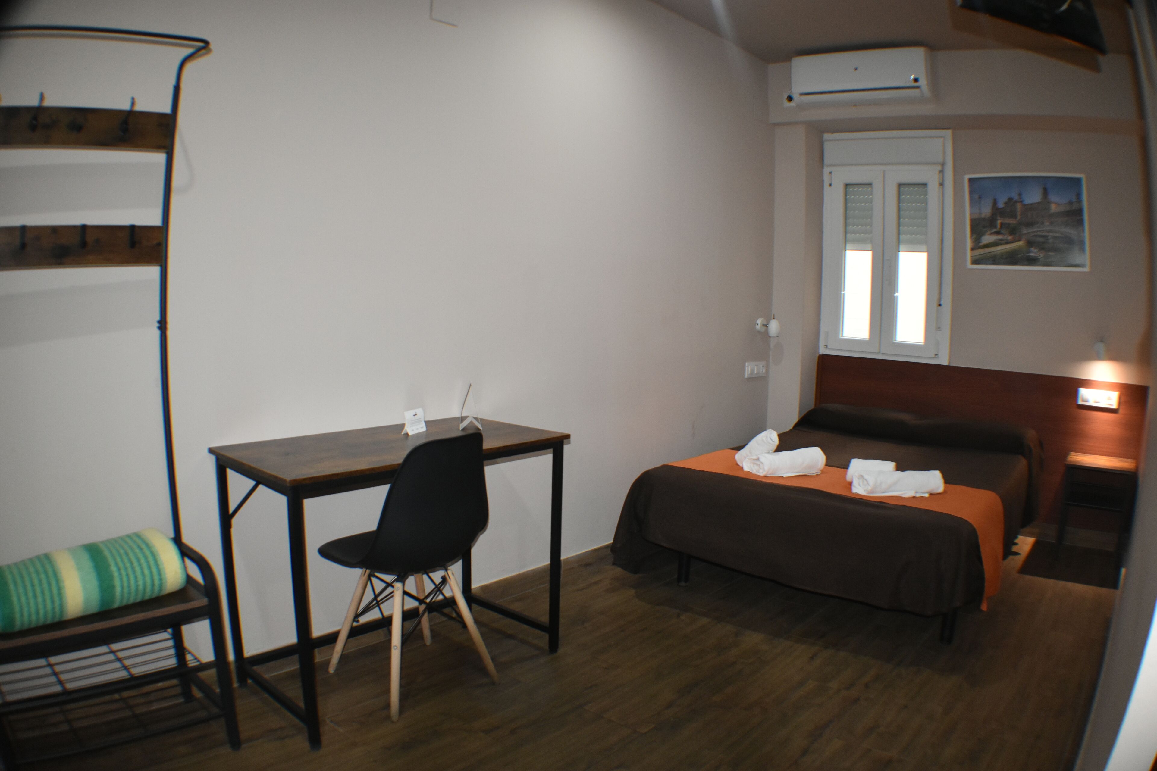 A compact hotel room with a single bed, a desk with a chair, and a small seating area with a bench.