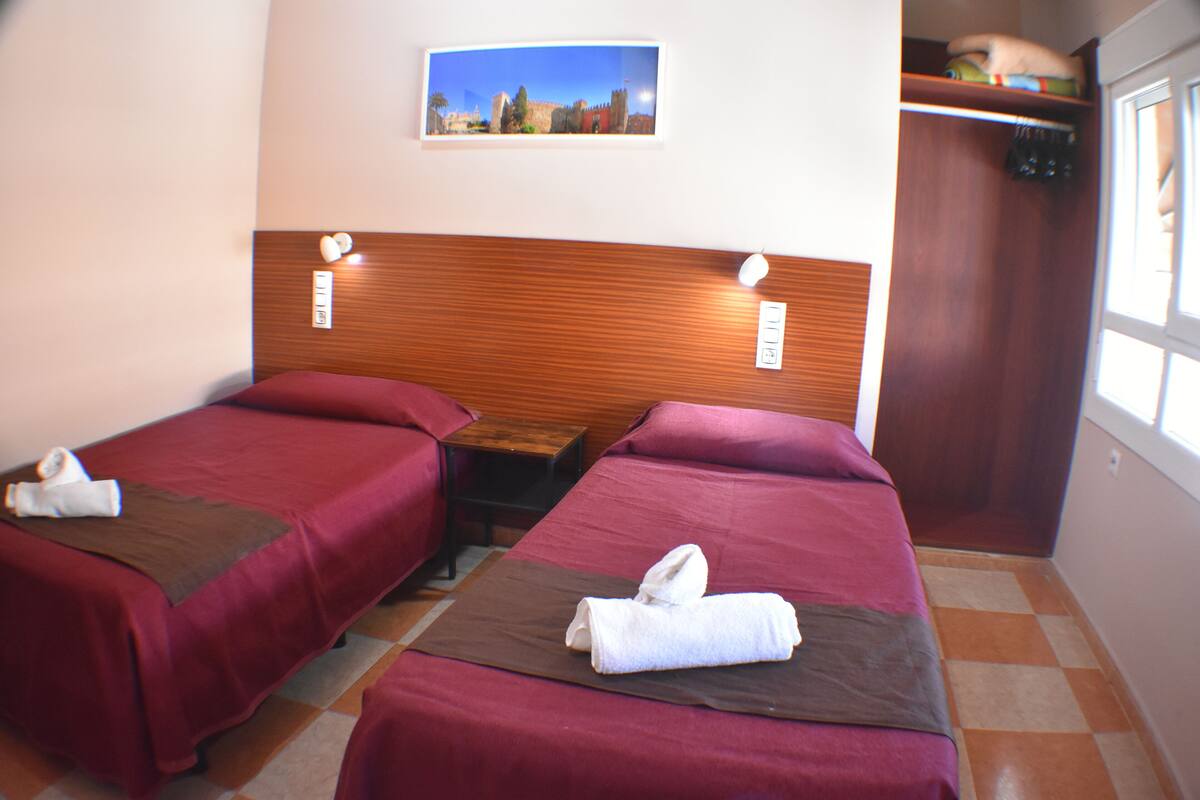 Hotellihuone, jossa on kaksi erillistä sänkyä, puiset päätyosat, pieni pöytä ja ikkuna.
