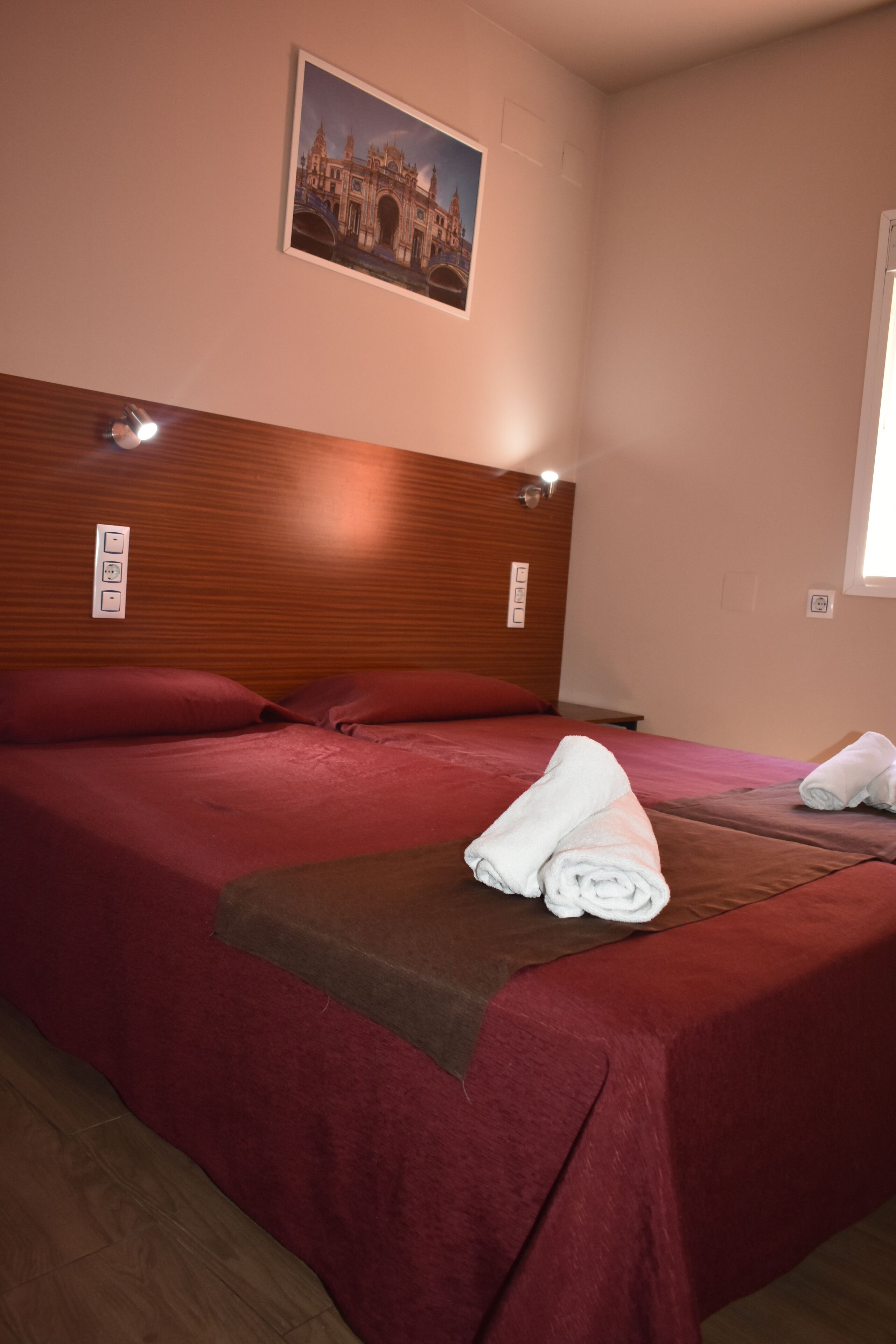 A hotel room with a wooden headboard, two beds with red bedding, a folded towel on the bed, and a framed picture of a bridge on the wall.