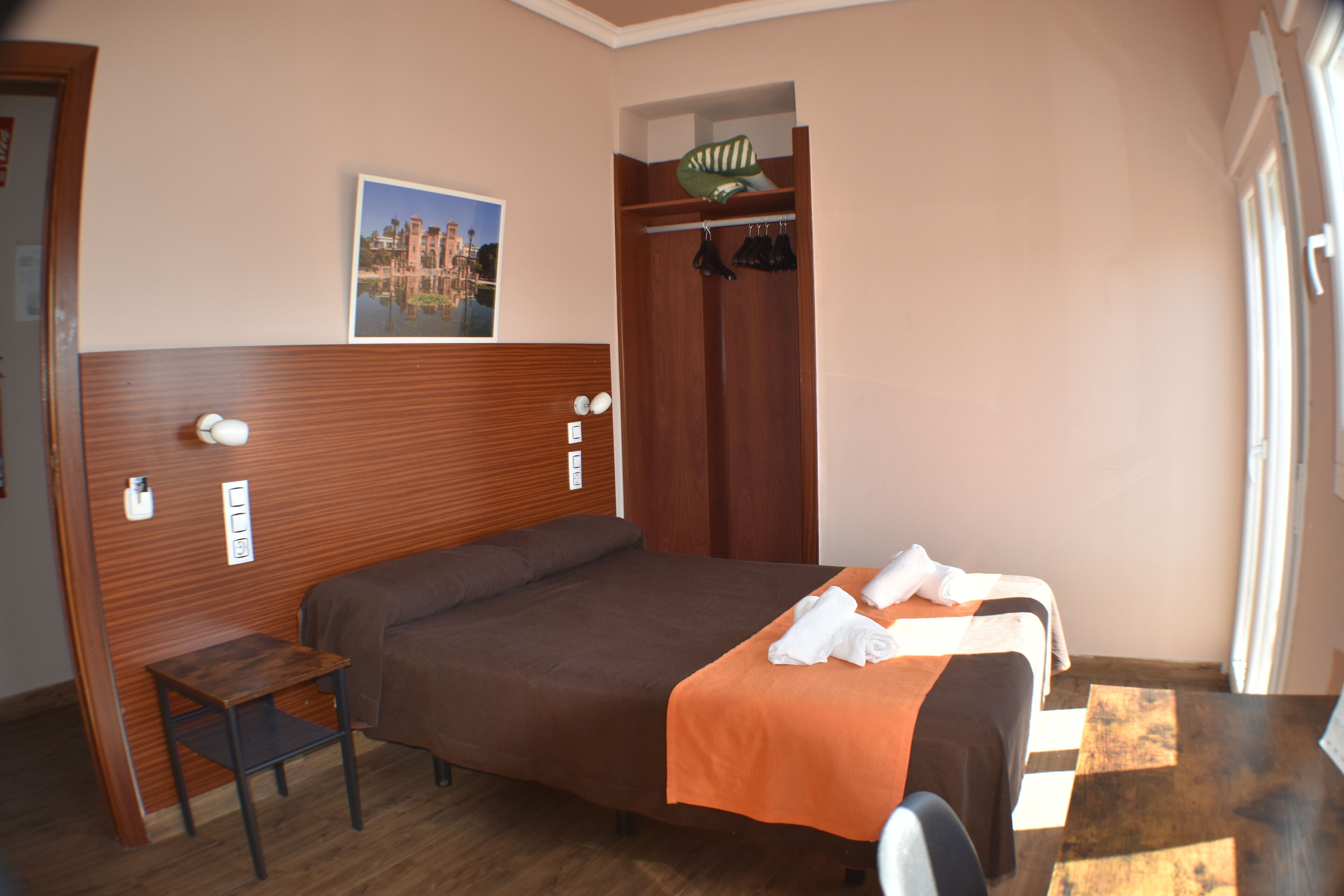 A hotel room with a wooden headboard, a bed with an orange bedspread, a small table, and a coat rack with clothes.