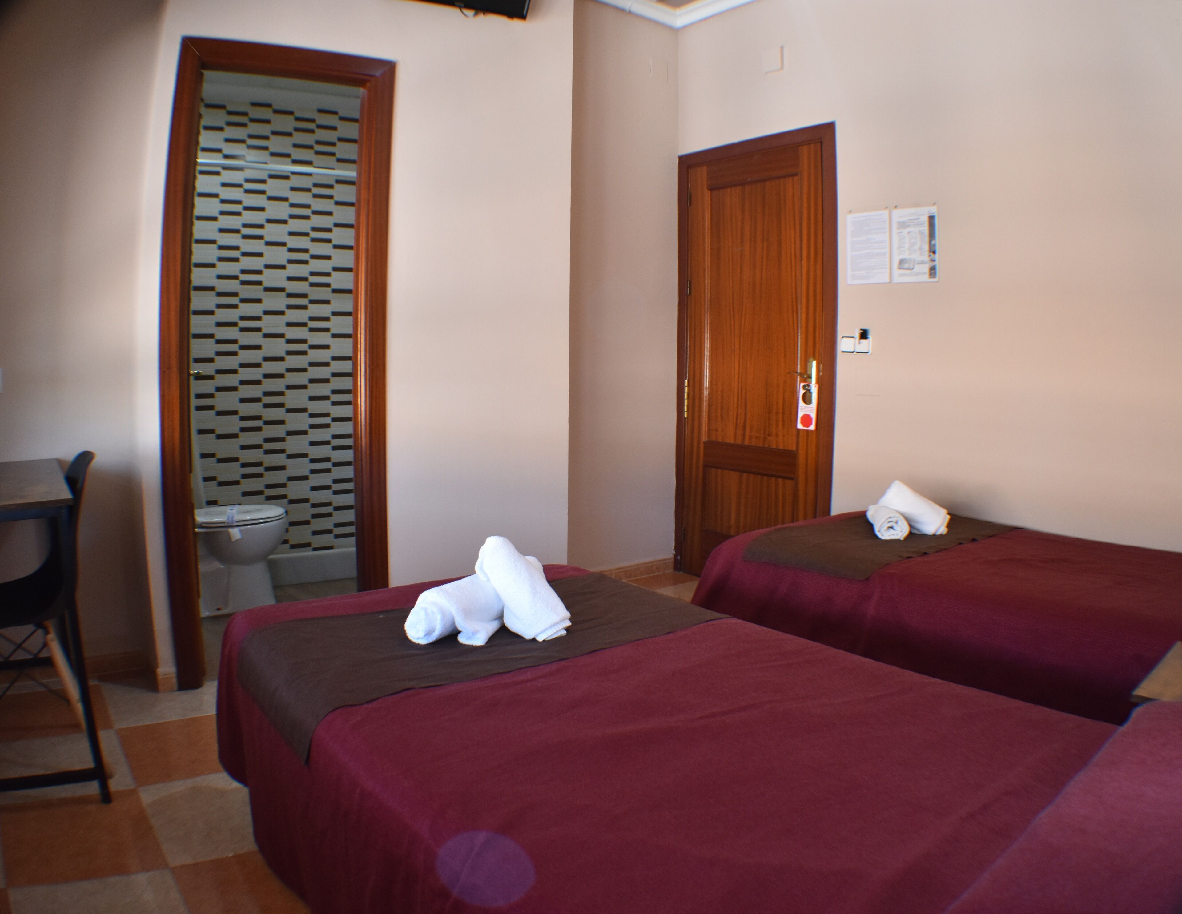 A hotel room with two beds, a wooden door, a bathroom with a patterned shower, and a small table.
