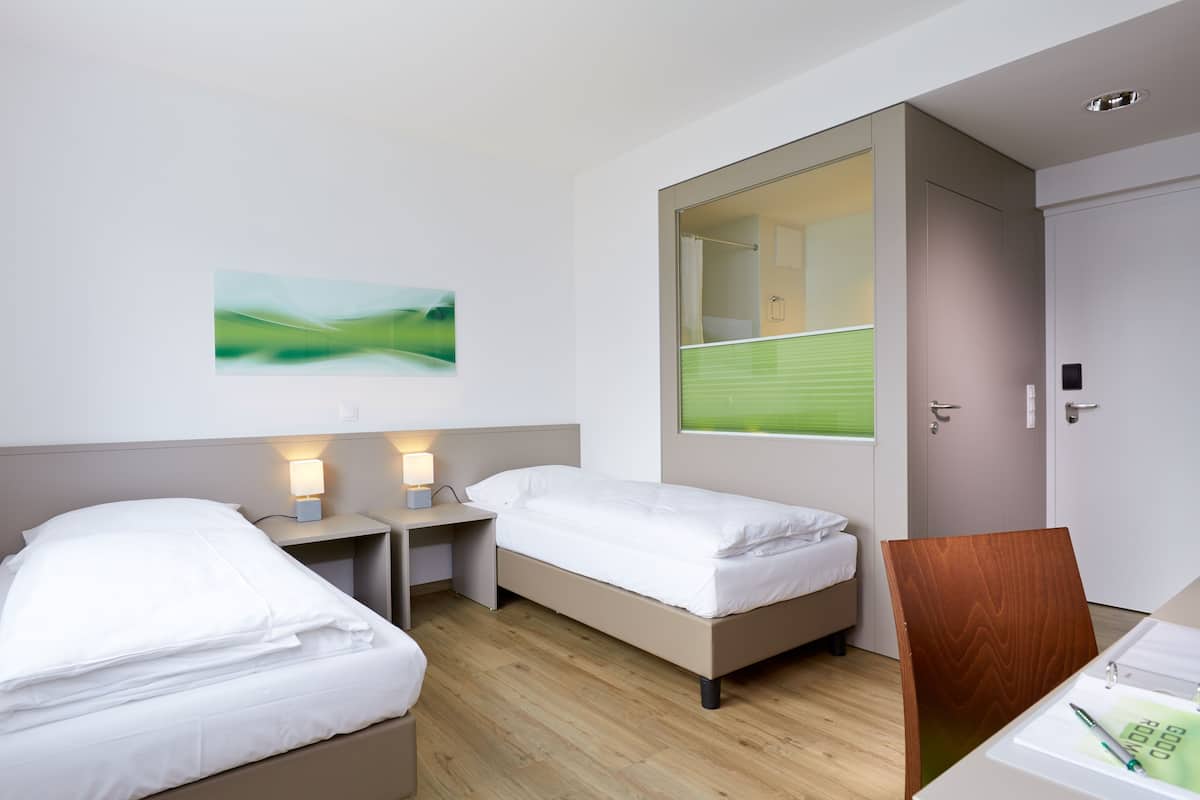 A hotel room with two single beds, a desk, a chair, a small table, a mirror, and a painting on the wall.