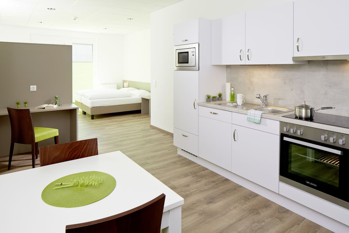 A modern kitchen with white cabinets, a microwave, and an oven. There is a dining area with a table and chairs, and a bedroom with a bed and window.