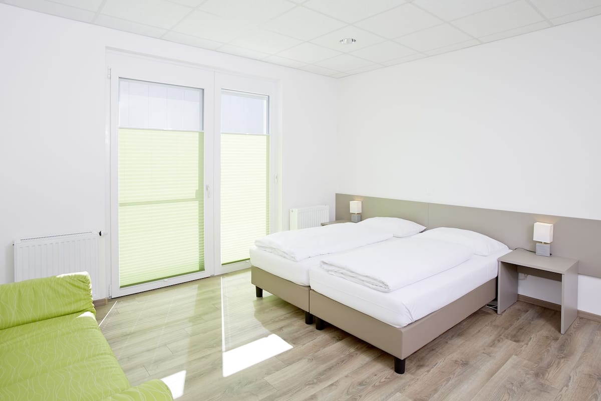 A modern bedroom with two beds, a green sofa, a desk, and a window with blinds.