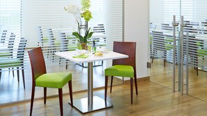 Un salón de conferencias con una mesa blanca, dos sillas verdes acolchadas, una planta en maceta y un panel de vidrio.