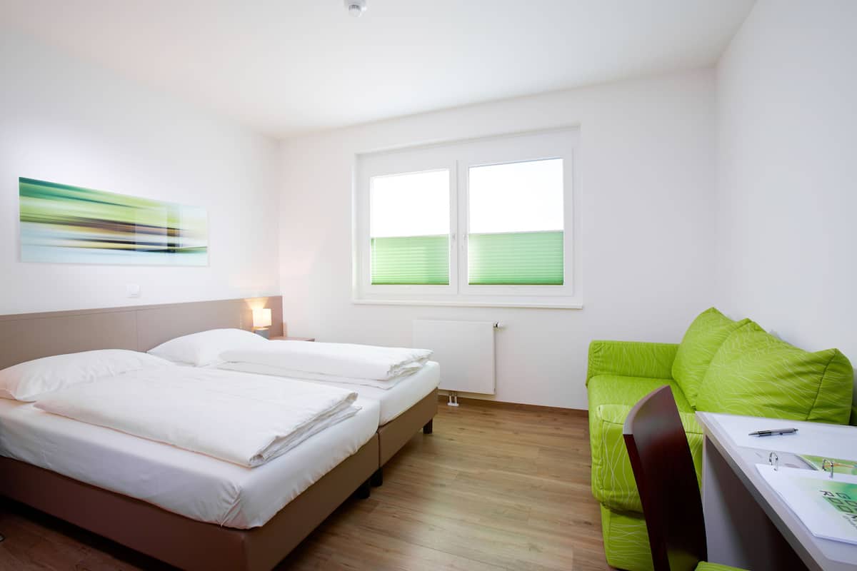 A modern hotel room with two beds, a green sofa, a desk with a chair, and a window with blinds.