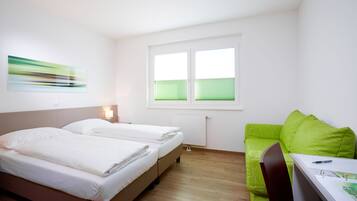 Ein modernes Hotelzimmer mit zwei Betten, einem grünen Sofa, einem Schreibtisch mit Stuhl und einem Fenster mit Jalousien.