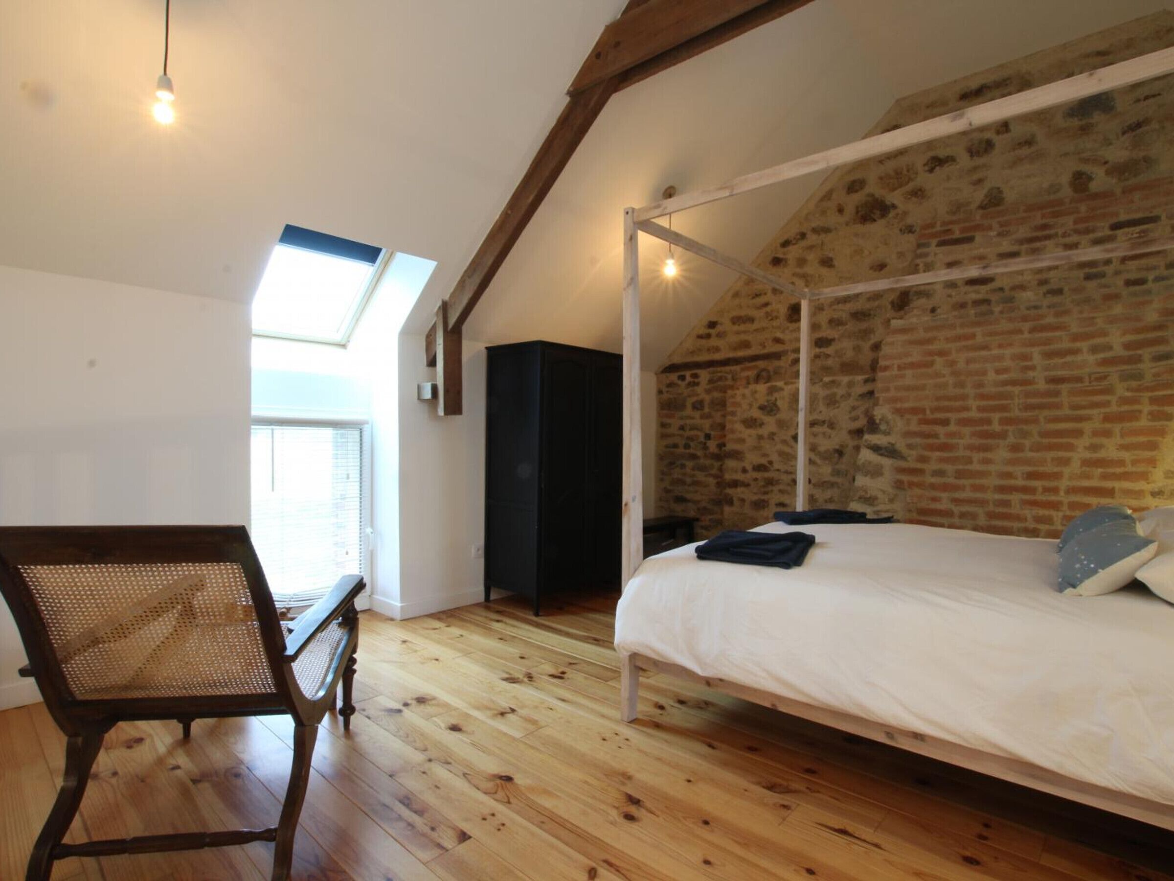 A bedroom with a sloped ceiling, exposed brick walls, a wooden floor, a four-poster bed, a chair, and a skylight.