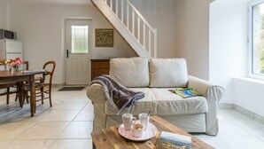 A cozy living room with a sofa, a coffee table with cups and a book, a dining area with a table and chairs, and a staircase leading to another level. - Charming Normandy cottage with wood stove and private yard (Bourguenolles)