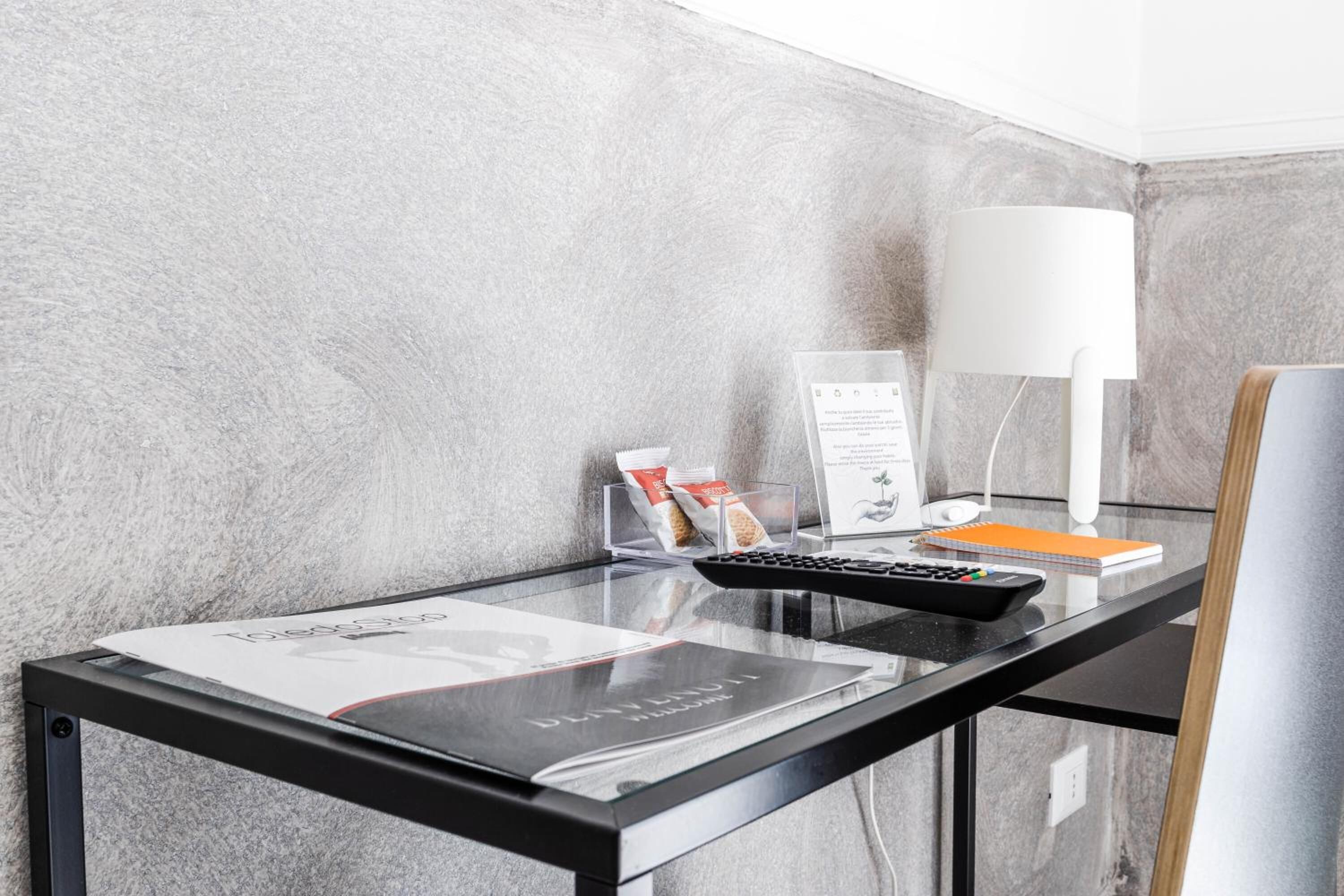 A modern hotel room with a glass desk, a remote control, a lamp, a notepad, and a magazine.