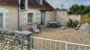 Une maison avec un toit en tuiles, un mur en pierre et une cour en gravier. Il y a une table et des chaises à l’extérieur.