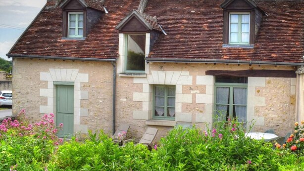 Une maison à deux étages, dotée d’un toit à deux versants, de nombreuses fenêtres et d’un jardin où poussent diverses plantes et fleurs.