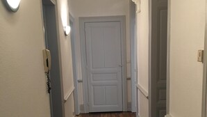 Un pasillo estrecho con suelo de madera y paredes blancas. Al final del pasillo hay dos puertas, una de ellas con un interruptor de luz arriba.
