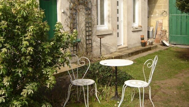 A rustic building with a green door, a small garden with two metal chairs and a table, and a ladder leaning against the wall.