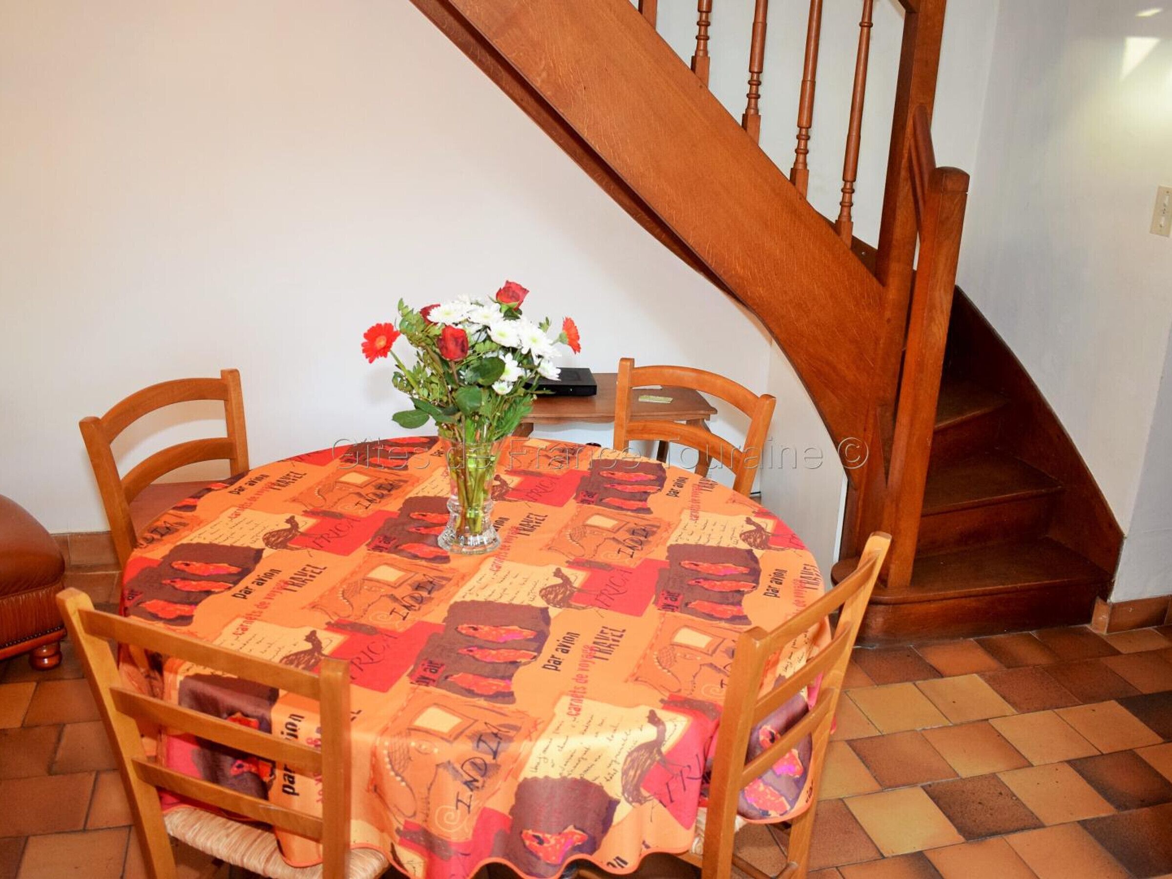Ein Essbereich mit einem runden Tisch, bedeckt mit einer gemusterten Tischdecke, umgeben von Holzsesseln und einer Vase mit Blumen.