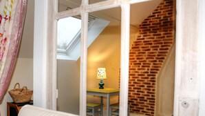 Una habitación con una silla blanca, una mesita con una lámpara y una cesta de mimbre. Se ve una pared de ladrillo y una ventana con tragaluz al fondo.