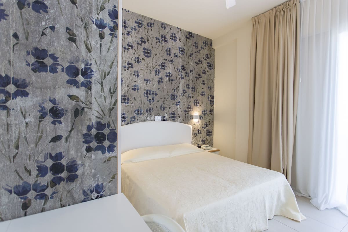 Une chambre à coucher avec un lit, une table de chevet, un mur recouvert d’un papier peint à motifs floraux et une fenêtre avec des rideaux.