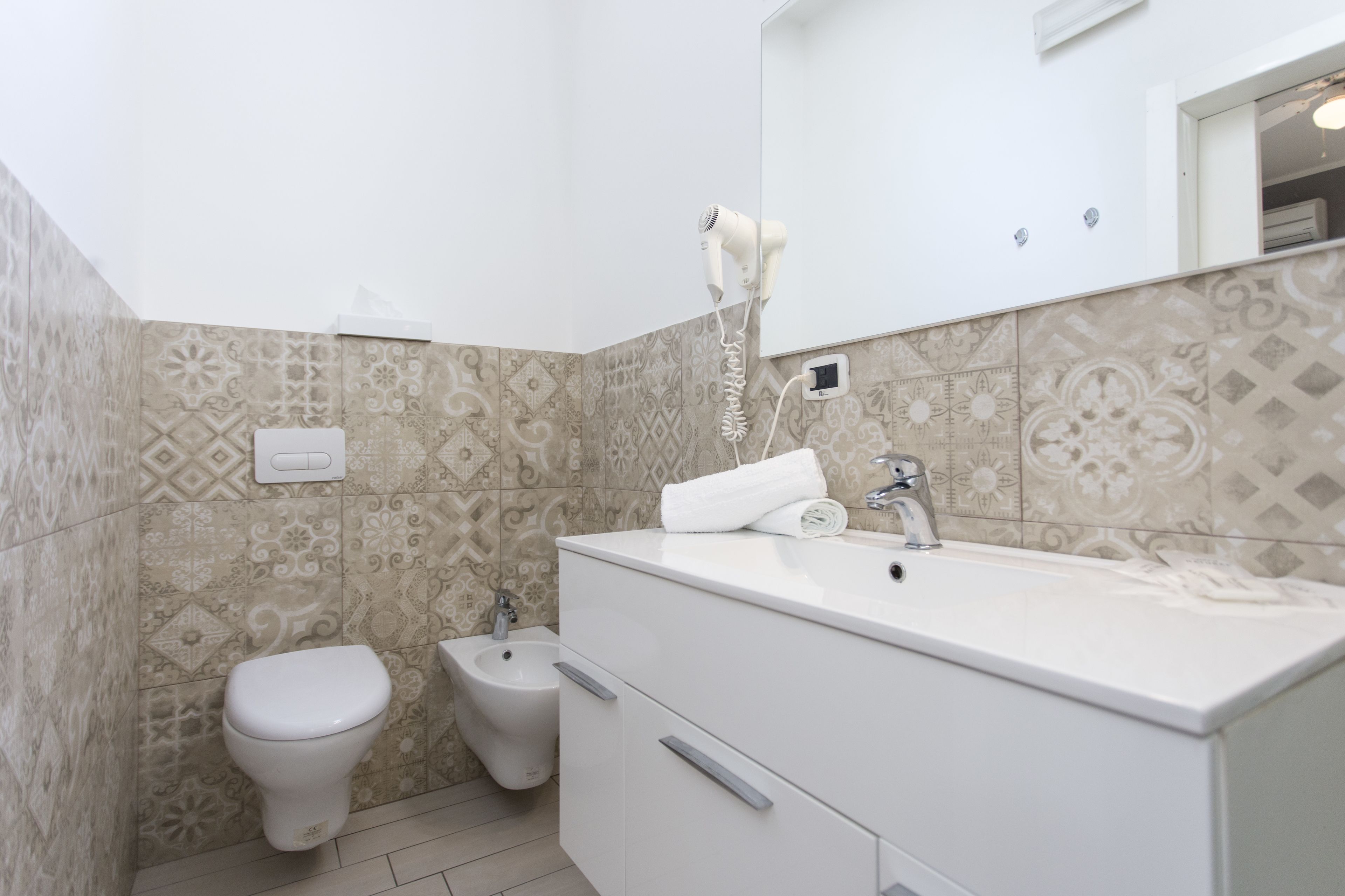 Une salle de bain équipée de toilettes, d’un bidet, d’un meuble-lavabo avec miroir et d’un sèche-cheveux fixé au mur.