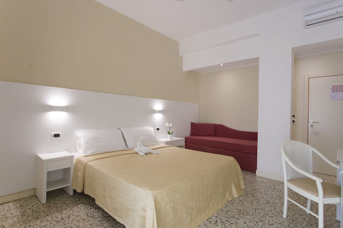 Une chambre d’hôtel avec un lit, une table de chevet, un canapé rouge, une chaise blanche et une porte.