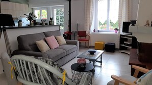 Una sala de estar con un sofá gris, una mesita, un mueble para televisión y una silla. Hay una ventana con cortinas y vistas a zonas verdes.